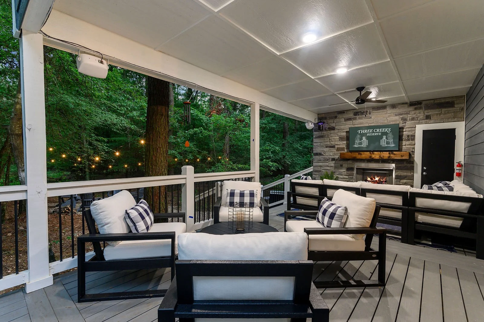 Outdoor covered patio with white and black furniture, string lights, a fireplace with a TV above it, and surrounded by tall trees.