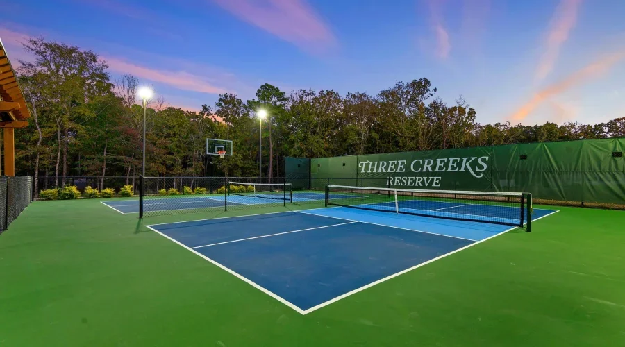 Two tennis courts at Three Creeks Reserve during sunset, with trees in the background and referee lamps illuminating the courts.