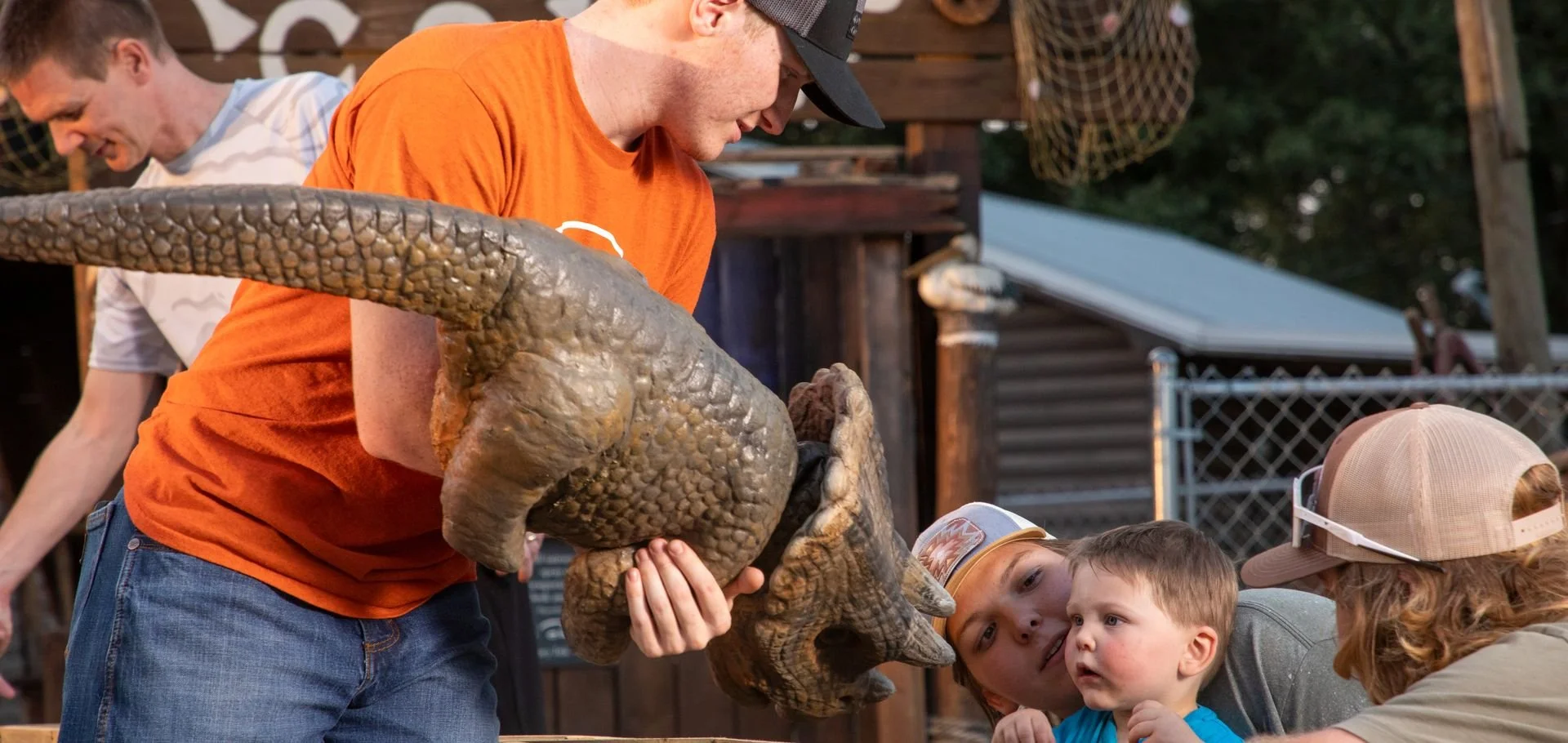 People interact with a small dinosaur prop during an outdoor event, with children observing and a man in an orange shirt holding the dinosaur.