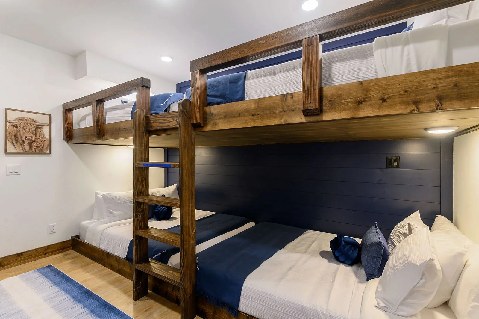 Bedroom with a wooden bunk bed resting above two beds with white and blue bedding. There is a framed animal portrait on the white wall.
