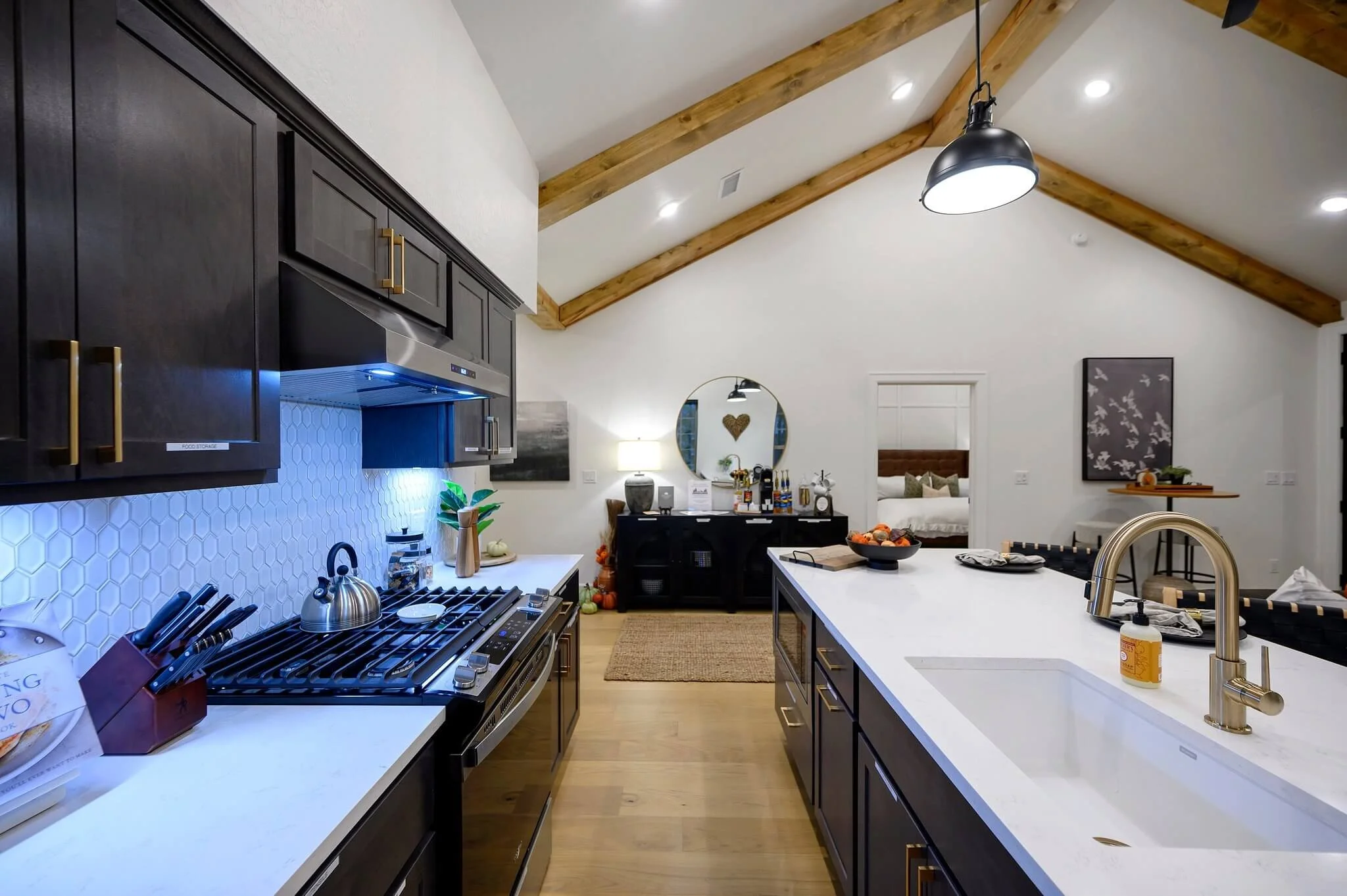 Modern kitchen with white countertops, dark cabinets with gold handles, and a vaulted ceiling with wooden beams. There is a gas stove with a stainless steel kettle, and a kitchen island with a gold faucet. The background includes a dining area and a 