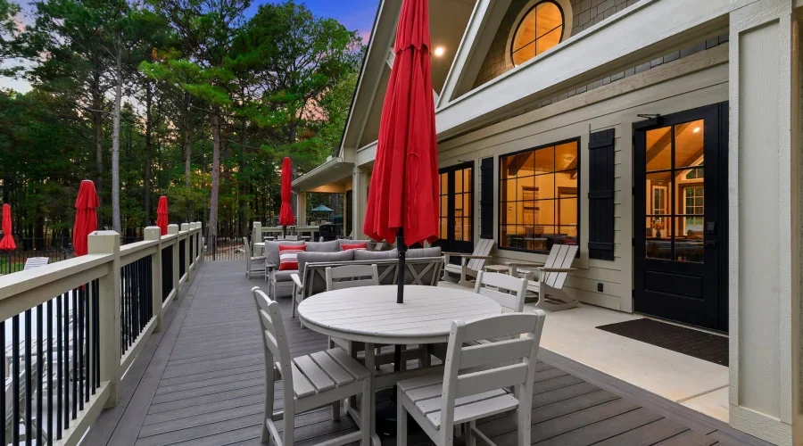 Outdoor patio area with white wooden furniture, red umbrellas, and a house with large windows and black shutters, surrounded by trees.