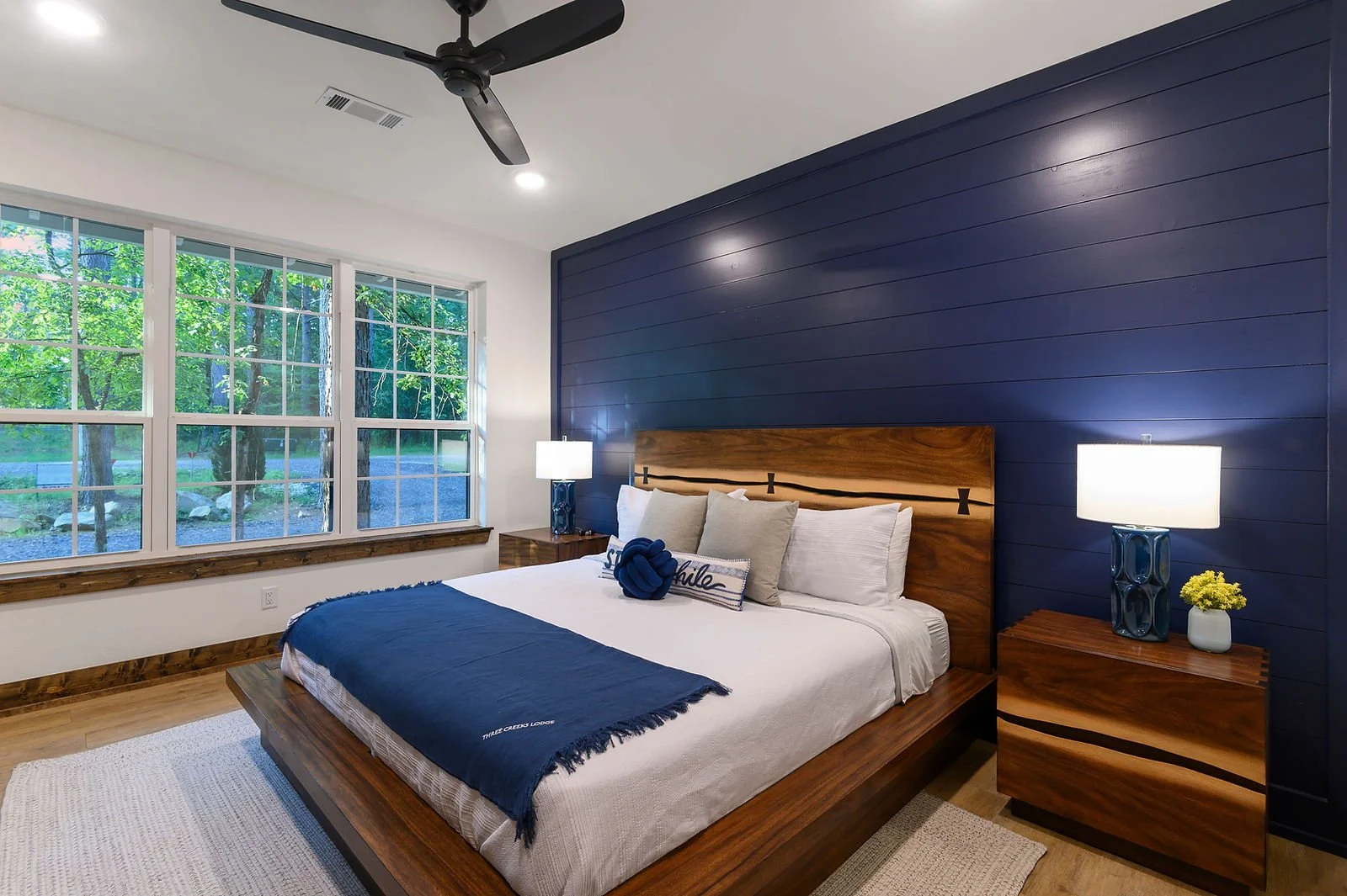 A modern bedroom with a large bed, navy blue accent wall, wooden headboard, nightstands with lamps, and a window showing green trees outside.