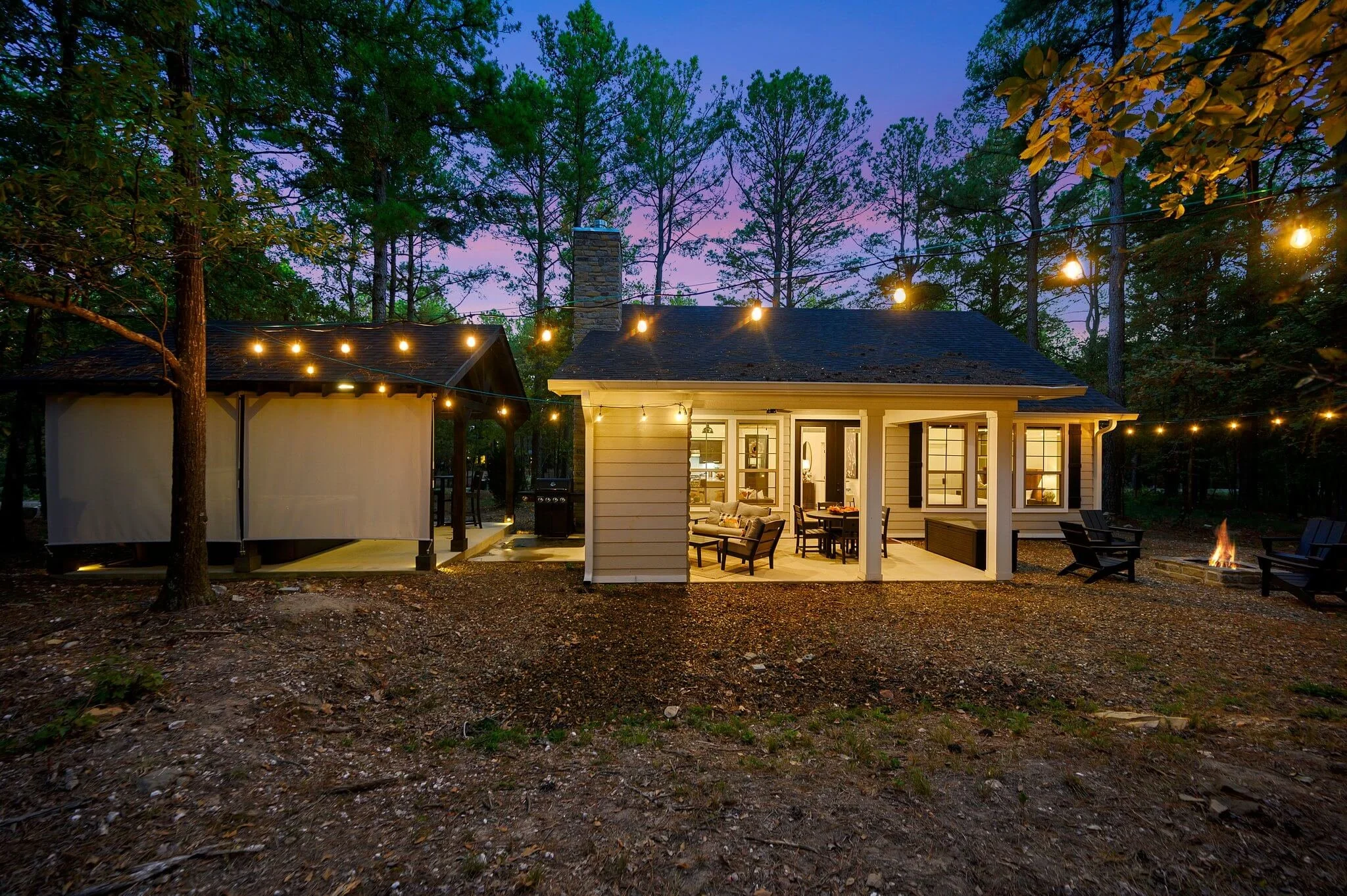 A backyard evening scene with a house, outdoor furniture, string lights, and trees.
