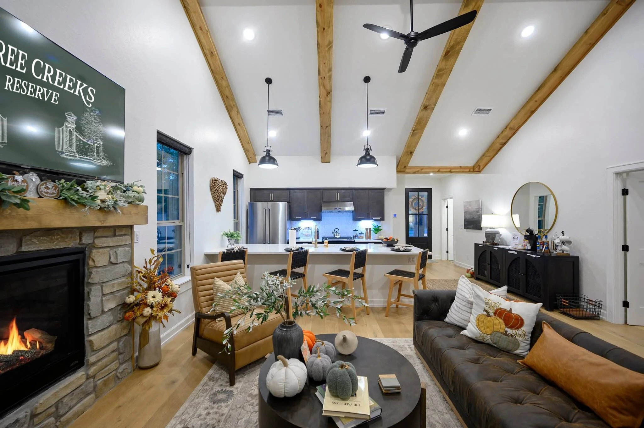 Modern open-concept living room and kitchen with wooden ceiling beams, a ceiling fan, a stone fireplace, decorative pumpkins, and fall-themed decor.