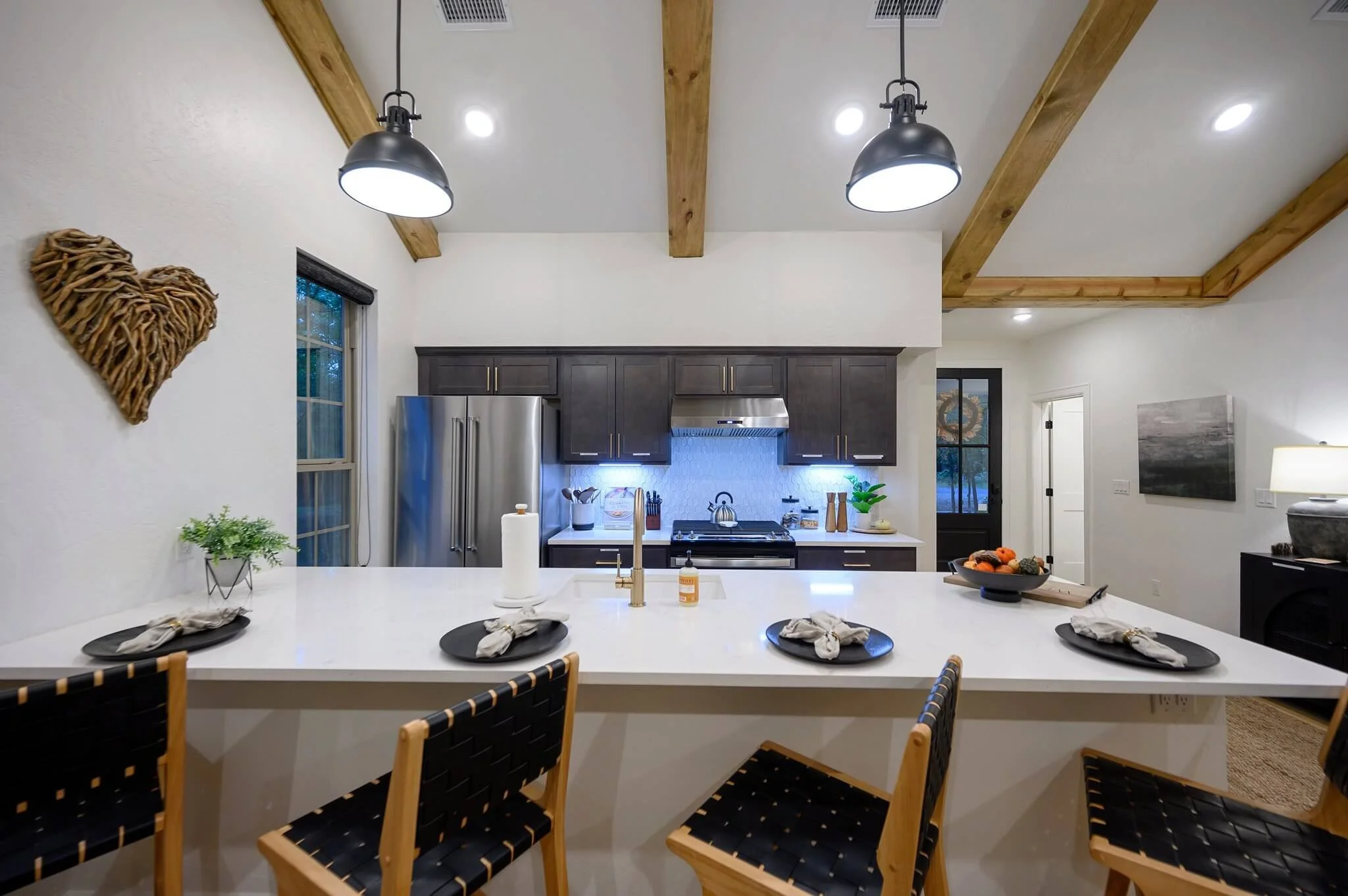 Modern kitchen with dark wood cabinets, stainless steel refrigerator, and a white island counter with black plates, napkins, and utensils. Wooden beams on the ceiling and decorative heart-shaped wall art. Small plants and kitchen accessories are on t