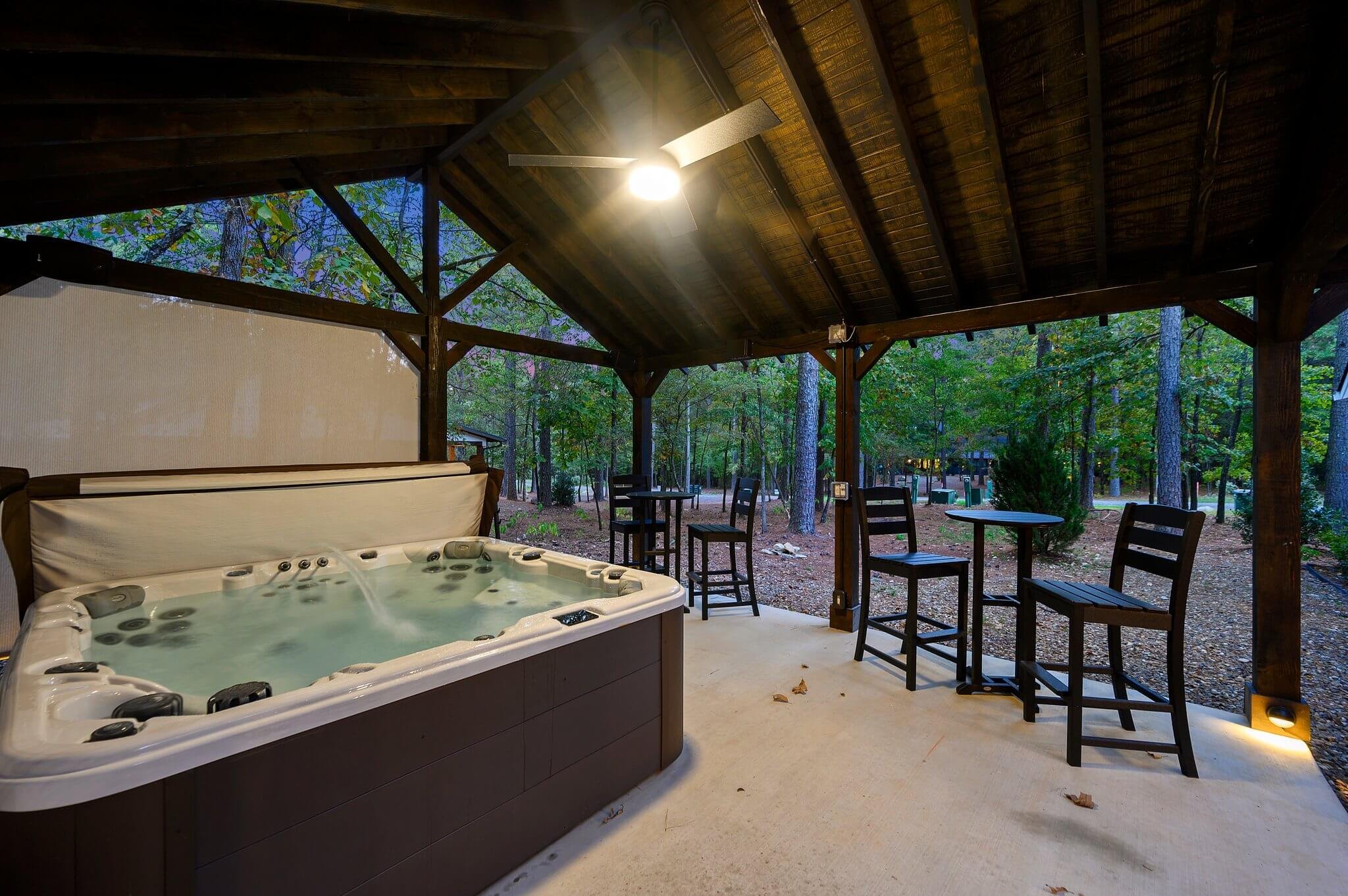 Outdoor patio area with a hot tub, high chairs, and tables, surrounded by trees and a wooden roof.