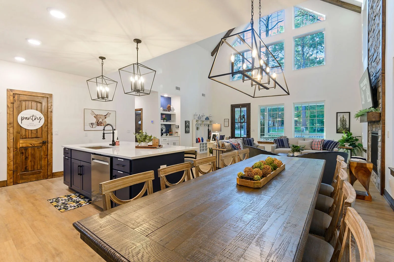 Spacious open-concept living and dining area with large windows, high vaulted ceiling, modern black kitchen island, wooden dining table with chairs, cozy seating with patterned pillows, and decorative fall centerpiece.