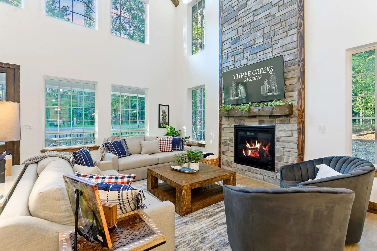 Living room with large windows, stone fireplace with a television on top, and cozy seating area including a sofa, armchair, and various pillows with a wooden coffee table and green plants.
