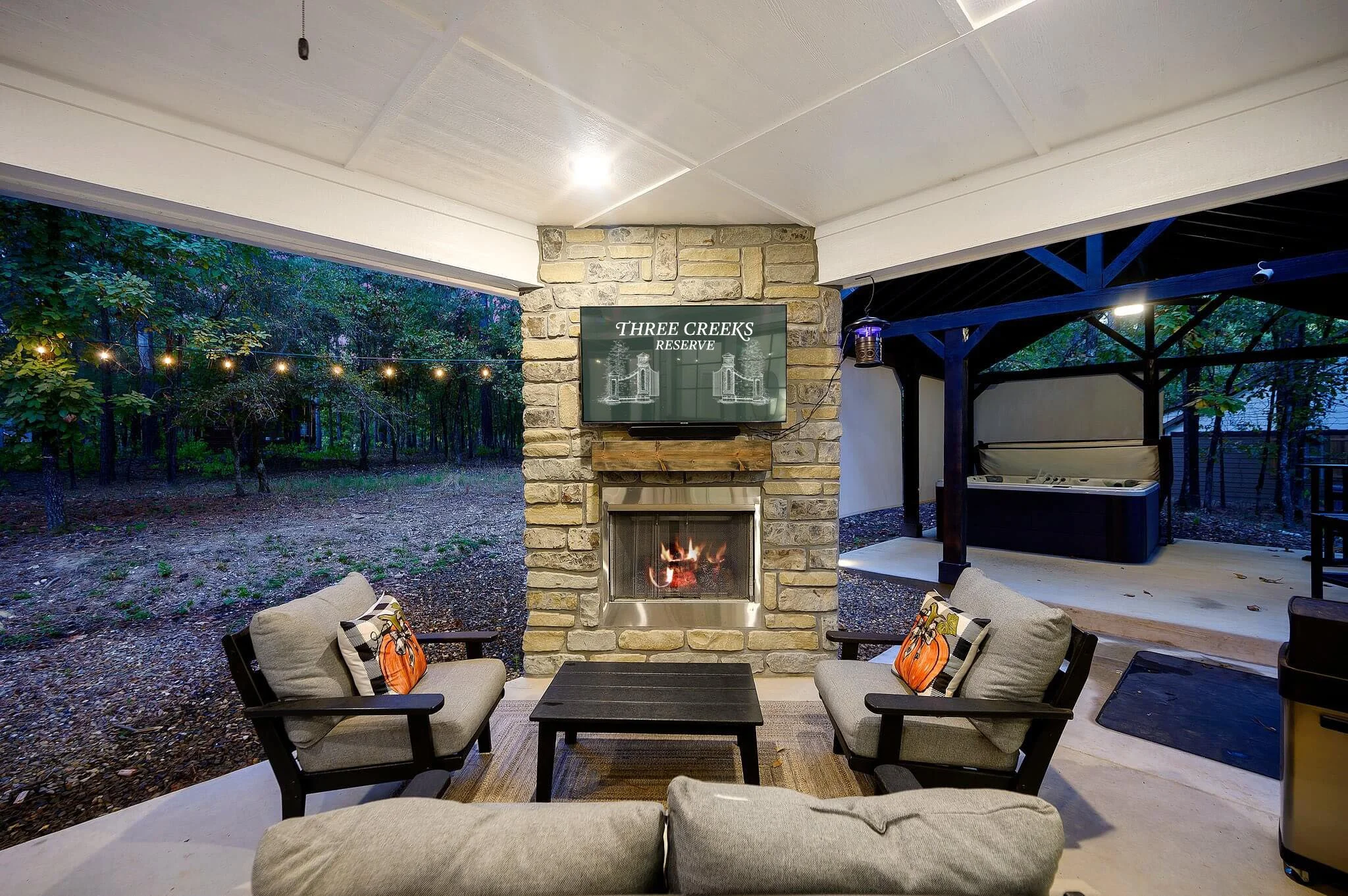 Outdoor patio with a fireplace, surrounded by seating with cushions and pillows, and a view of a wooded area with string lights.
