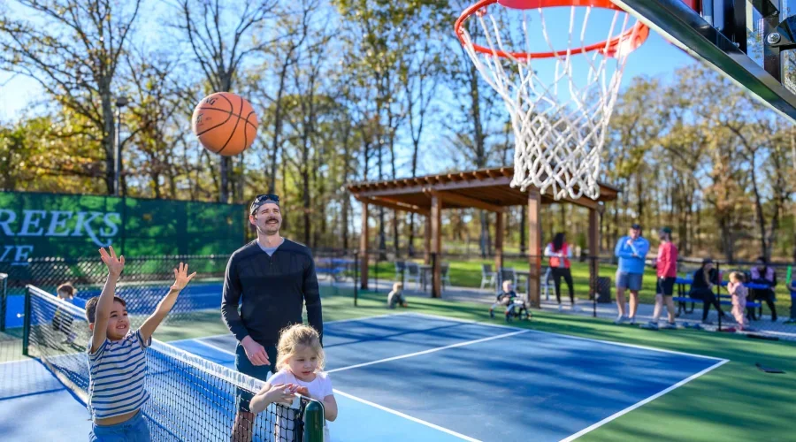 A man and two children playing on a public outdoor basketball court during daytime. The girl in the striped shirt is shooting a basketball while the boy in white watches. There are other people in the background, some sitting on benches and others wa