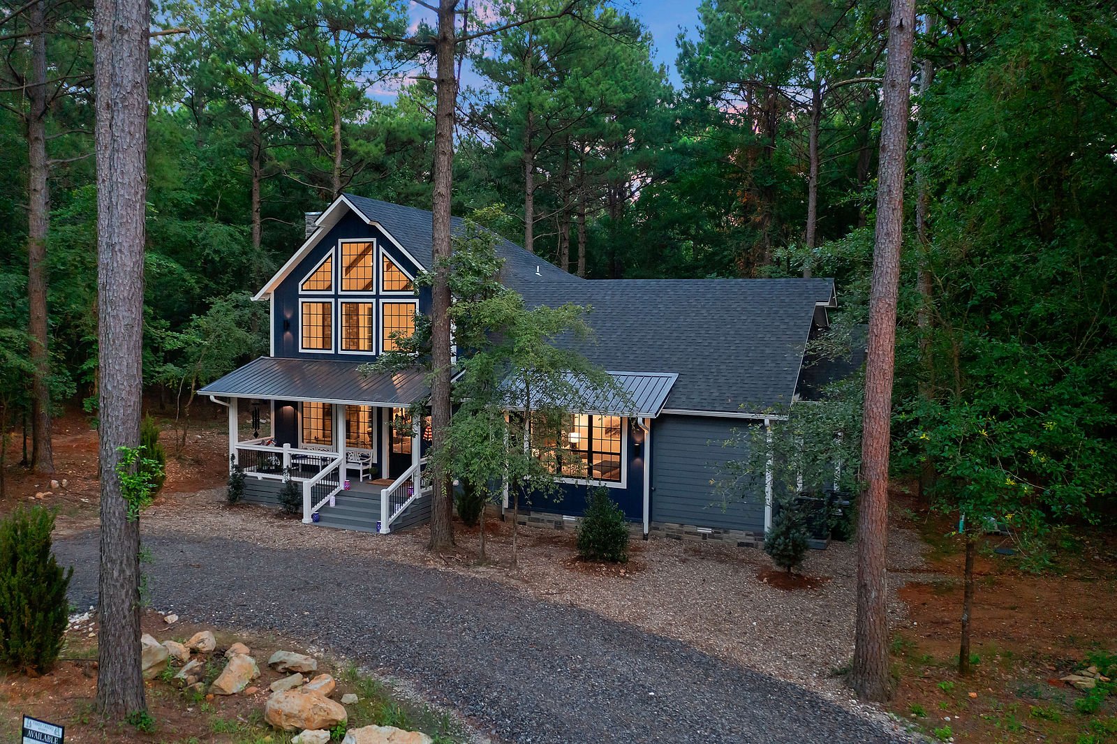 A two-story house with large windows, painted dark blue with white trim, is nestled among tall trees in a wooded area. The house features a front porch with stairs, a gray roof, and warm lighting inside. The setting appears to be at dusk or early eve
