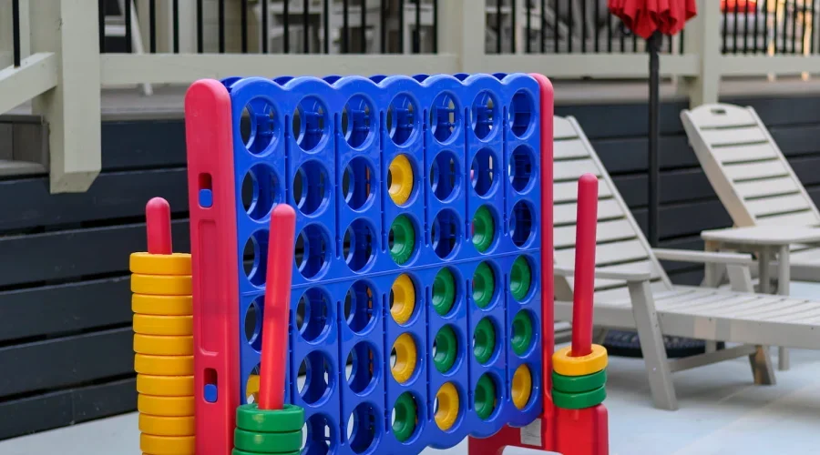 Large Connect Four game with colorful discs, set up outdoors on a deck with two lounge chairs and a patio umbrella in the background.