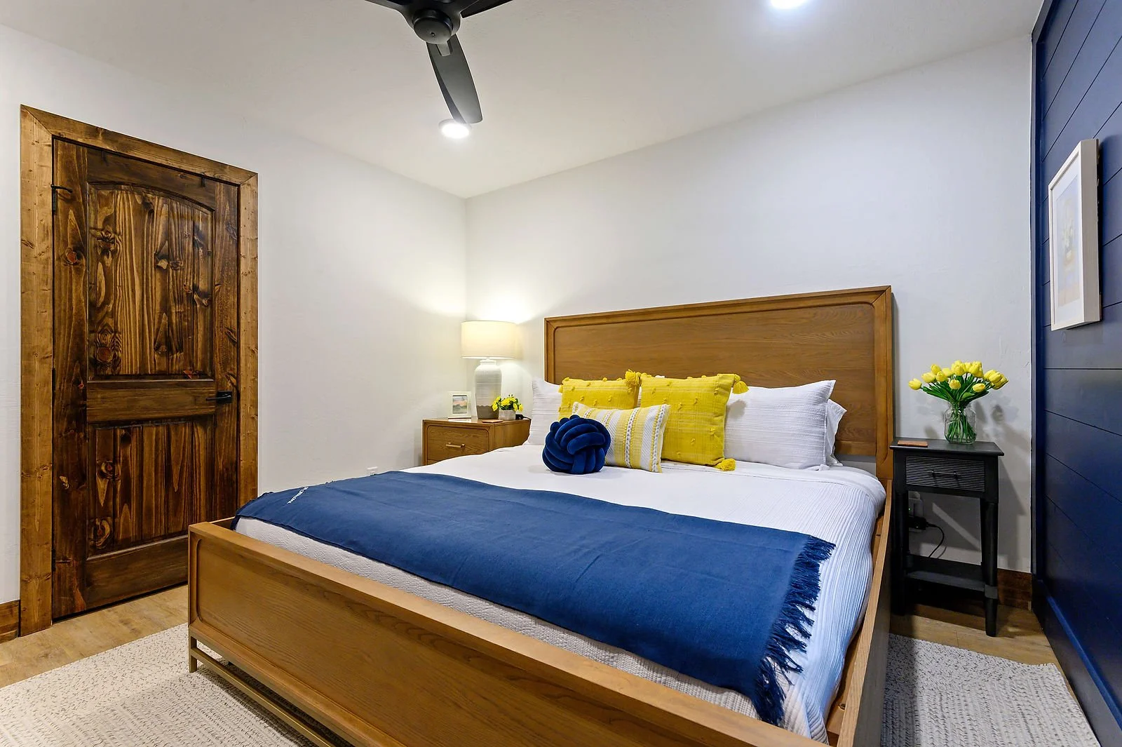 A bedroom with a wooden bed frame, white bedding, blue throw blanket, and yellow pillows. Two black nightstands with yellow flowers, a lamp, and a picture are on each side. A ceiling fan with a light is above, and a textured light-colored rug covers 