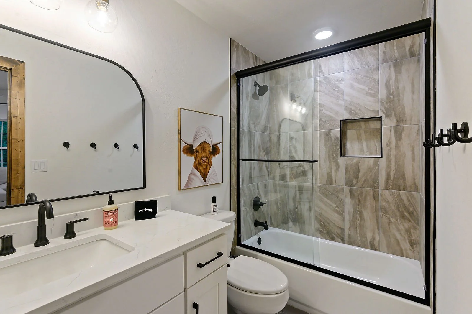 Modern bathroom with white vanity, black fixtures, a large mirror, a framed cow artwork, and a walk-in shower with beige stone tiles and black hardware.