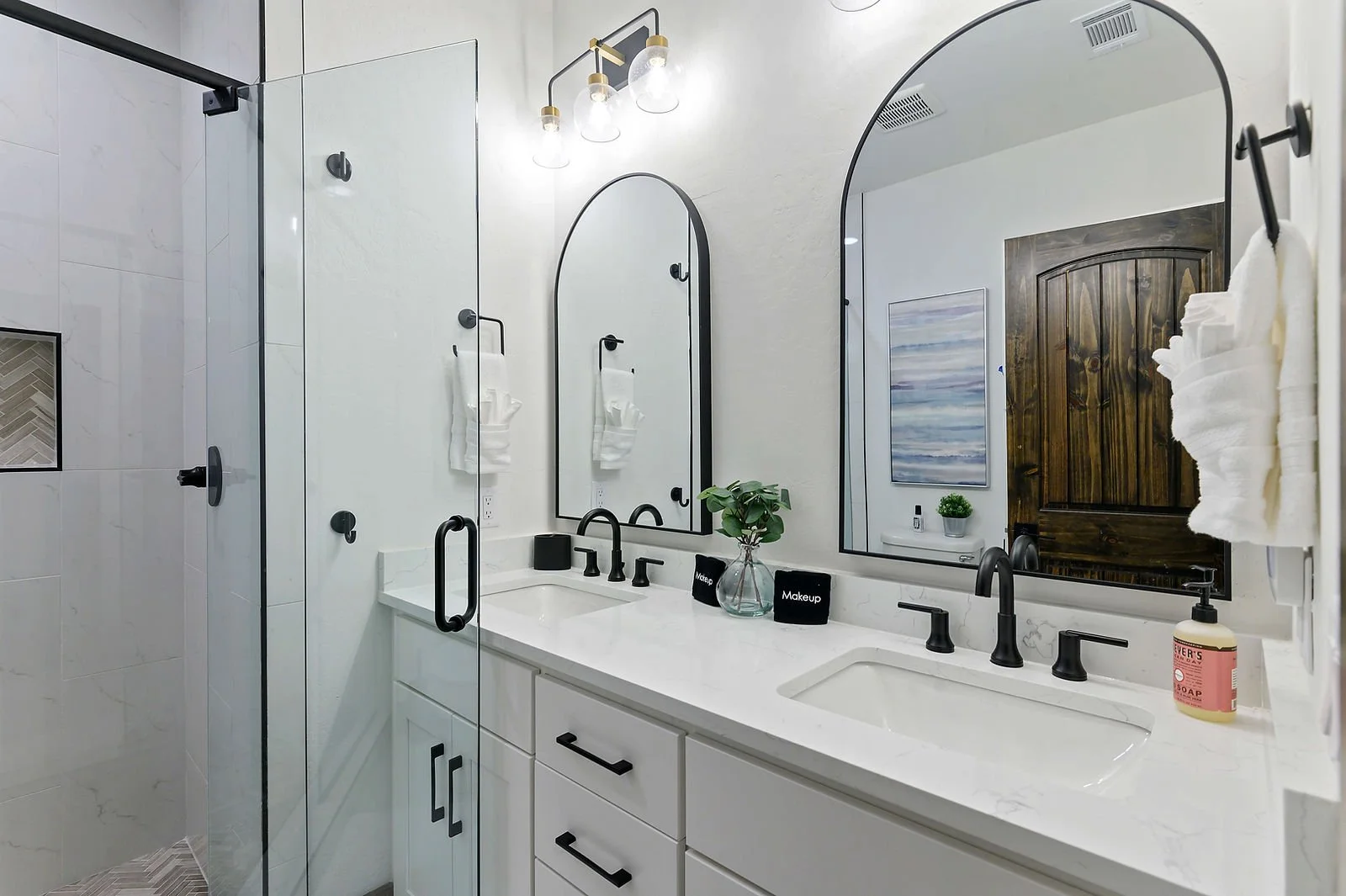 Modern bathroom with a white vanity, black fixtures, a large mirror, potted plant, soap dispenser, towels, and a glass shower enclosure with tiled walls.
