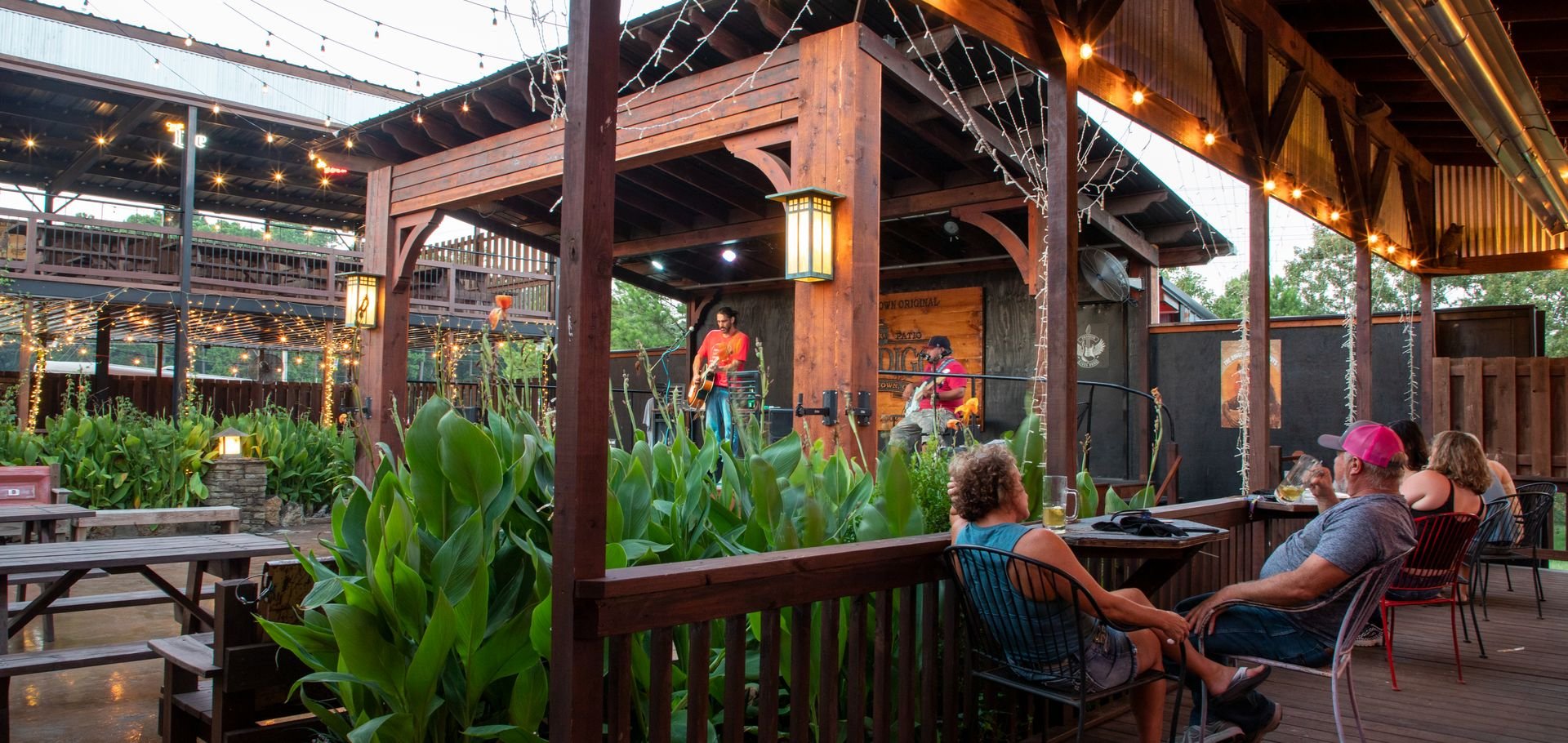 Musicians performing on an outdoor stage at a rustic restaurant or bar with string lights and greenery, while diners sit at tables in the foreground.
