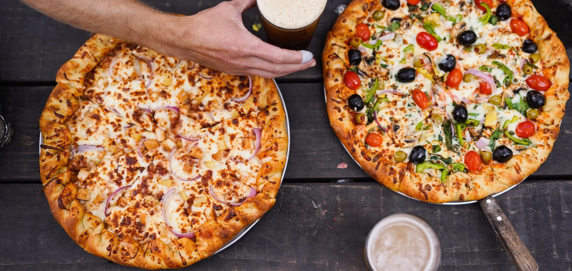 Two pizzas on a dark wooden table, one with cheese and onions, and the other with cheese, black olives, cherry tomatoes, green peppers, and onions, with two glasses of beer and a hand reaching for the beer.