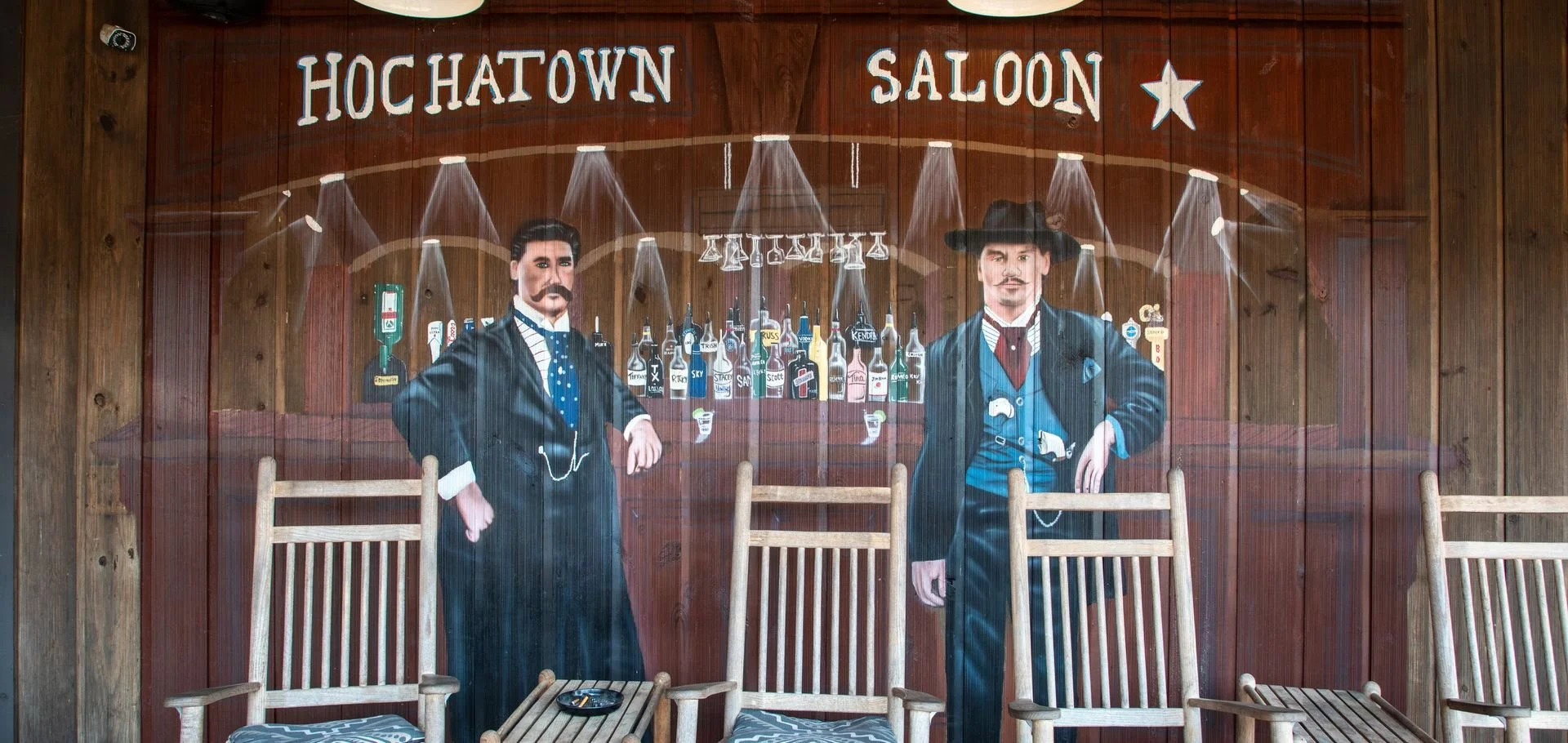 Mural painting of two men in old western attire standing in front of a bar with various bottles, in a saloon named 'Hochatown Saloon'.