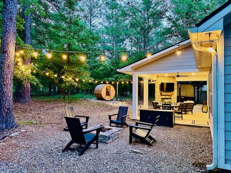Backyard with outdoor seating, string lights, a small covered patio with a fireplace and TV, a hot tub, and a wooded background.
