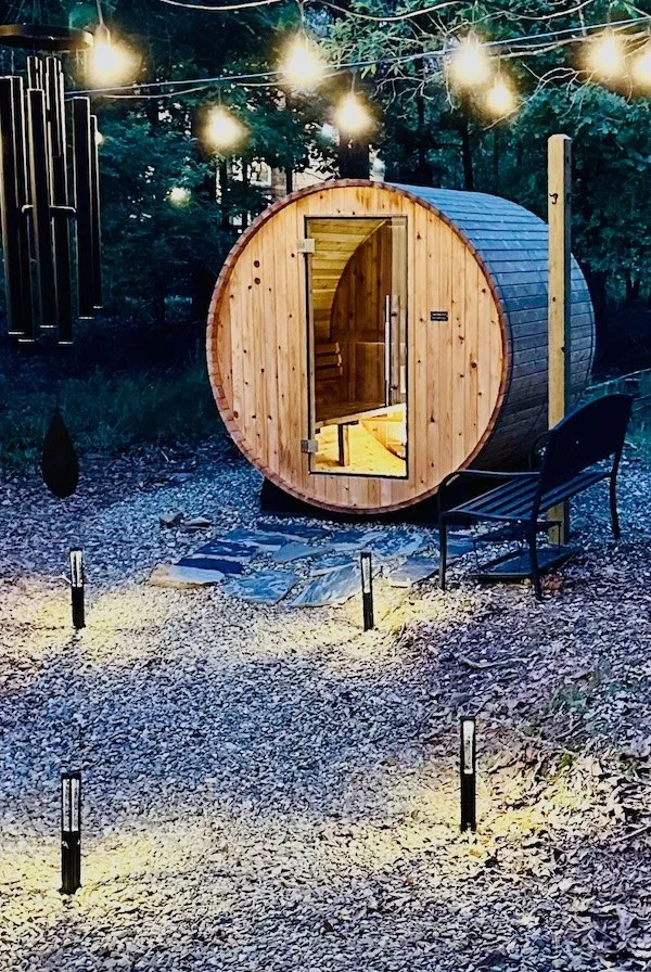 A small barrel-shaped outdoor sauna with a wooden exterior, string lights overhead, a black bench outside, and a stone pathway leading to the door in a wooded area.