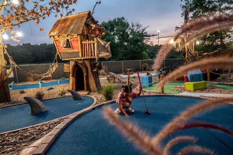 Child playing mini-golf at a whimsical outdoor playground with a pirate ship-themed structure and dolphin sculptures, surrounded by trees and a fence at dusk.