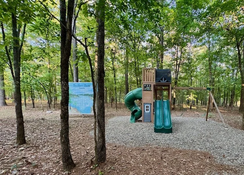 A playground with a treehouse structure, green slide, and a swing set surrounded by trees, with a sign that reads 'Broken Bow' nearby.