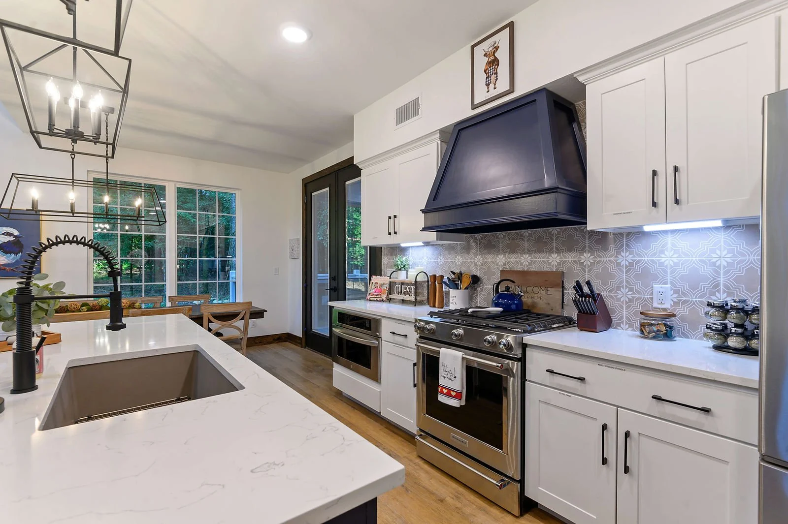 A modern kitchen with white cabinetry, stainless steel appliances, and a large island with a white marble countertop. There is a black range hood above the stove, decorative backsplash, and a dining area with wooden table and chairs near large window
