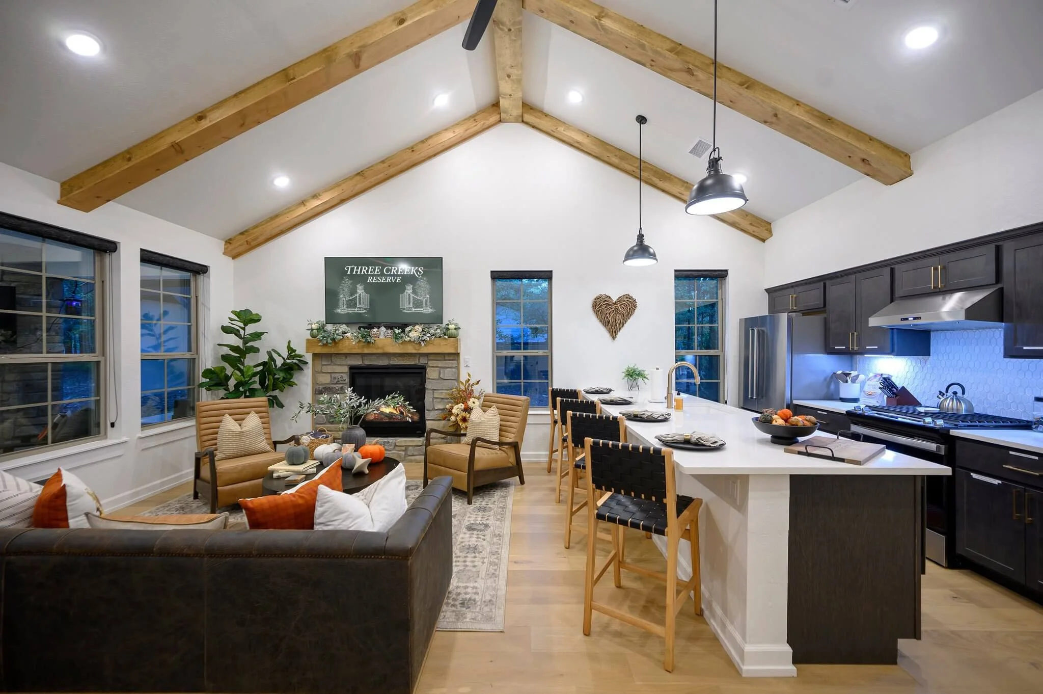 Open concept kitchen and living room with vaulted ceiling, wooden beams, and modern decor. The fireplace with a floral garland is on a wall with a TV above. There are windows with black frames, a dark-colored refrigerator, and a kitchen island with c
