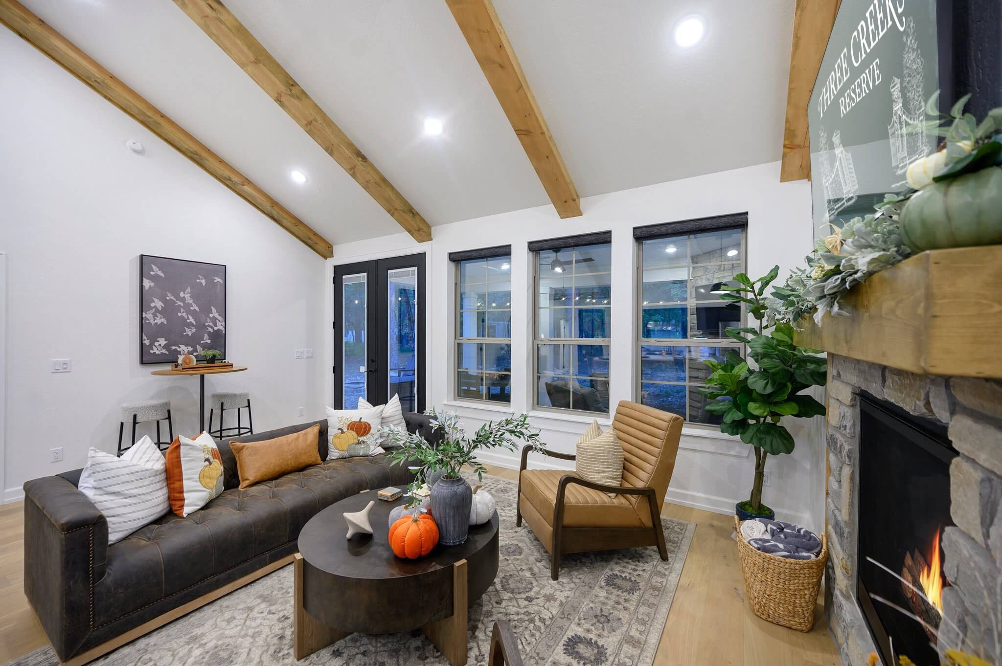 A living room with a vaulted ceiling with exposed wooden beams, white walls, and large windows. The room is decorated with pillows, plants, and wall art.