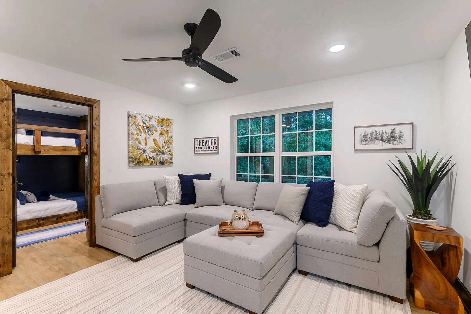 Living room with a white sectional sofa, blue and white pillows, wooden side table with a plant, artwork on the walls, large window showing trees, ceiling fan, and a glimpse of a bedroom with bunk beds.