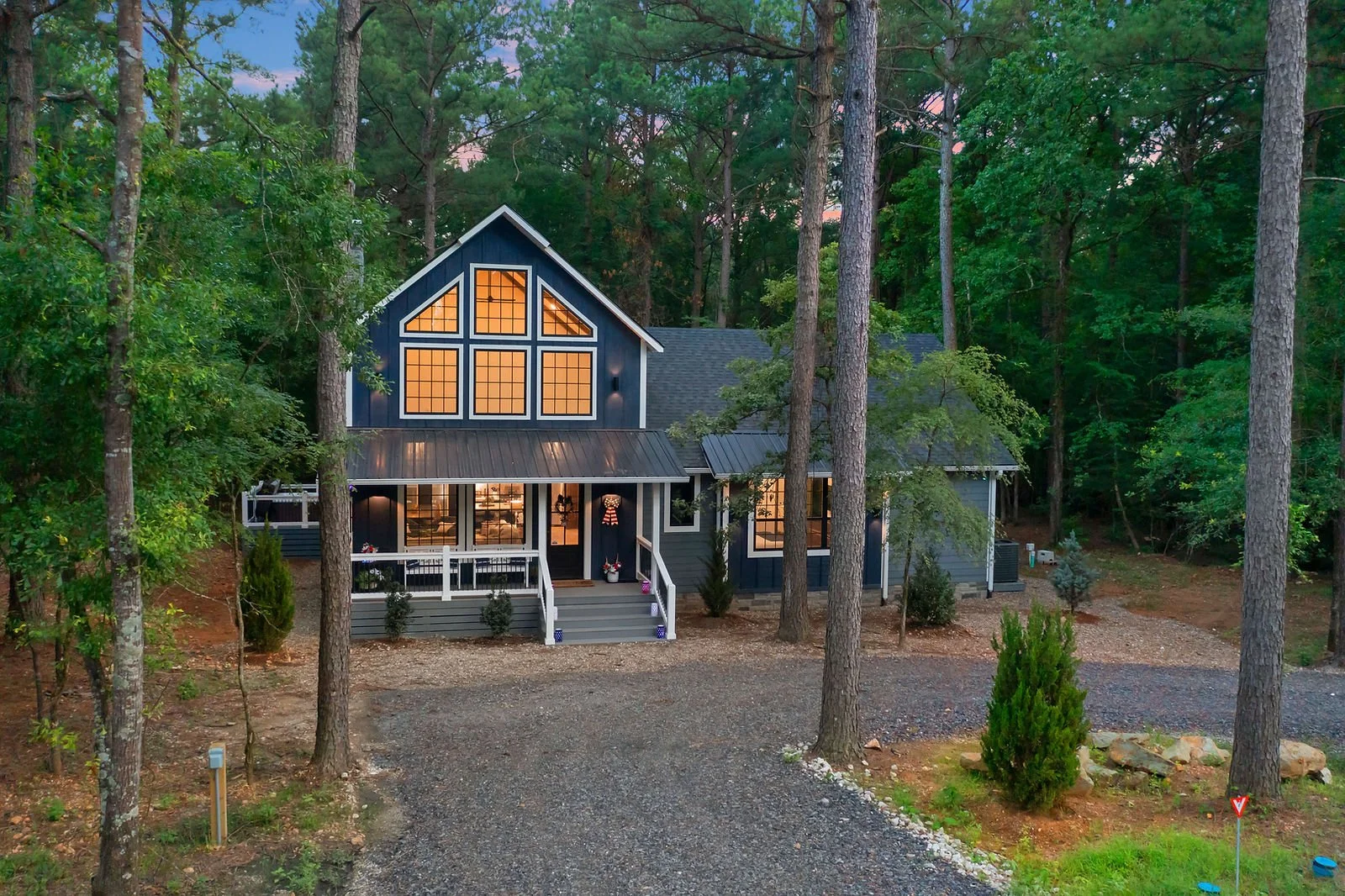 A modern house surrounded by tall trees at dusk, with warm interior lights visible through large windows, a gravel driveway leading to the front steps, and landscaped plants around the house.