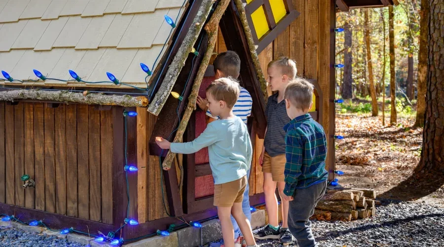 Group of children gathered outside a small wooden playhouse decorated with colorful string lights in a forest setting.
