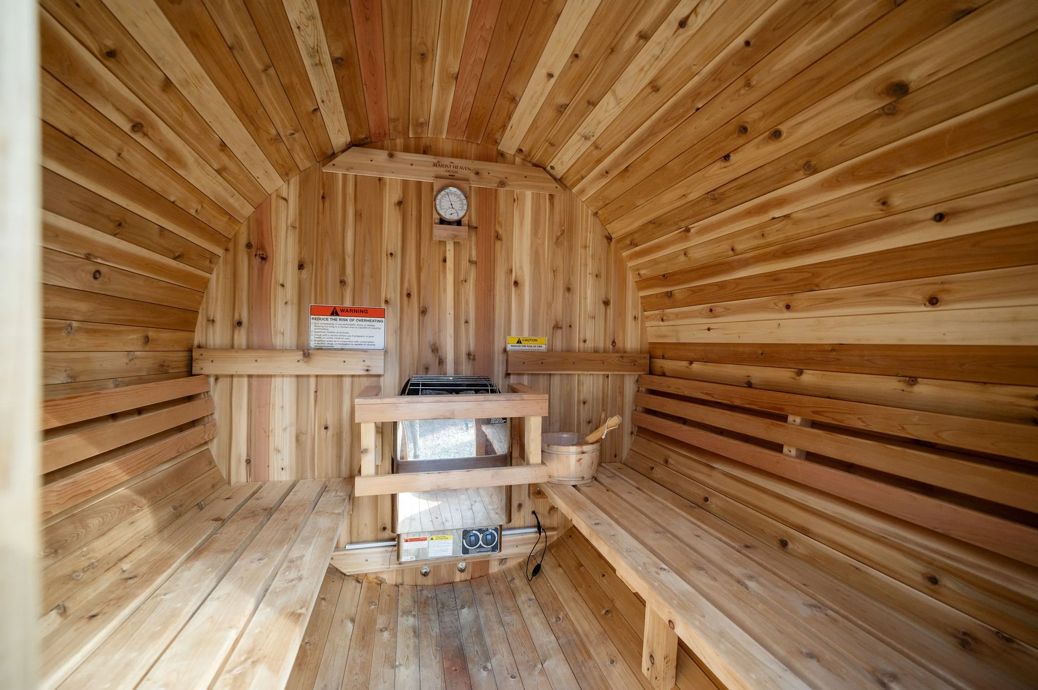Inside a small wooden sauna with wooden benches and a heater. A thermometer and warning signs are visible on the wall.