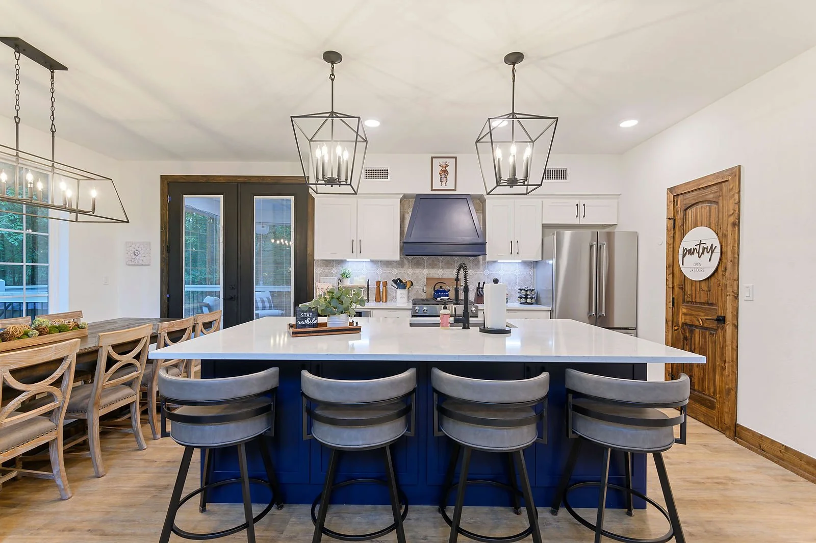 Modern kitchen with white countertops, gray and beige chairs, and wooden accents. Hanging light fixtures above the island, and a door with a round sign that says 'pantry open 24 hours'.
