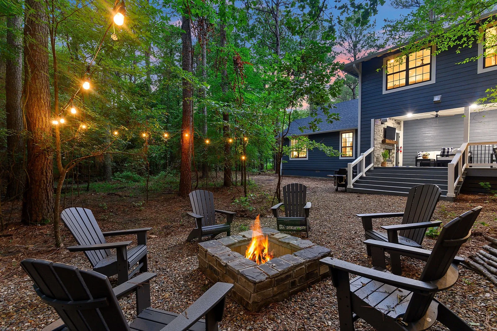 Outdoor backyard with a fire pit surrounded by Adirondack chairs, string lights hanging among trees, and a house with a screened porch illuminated at dusk.