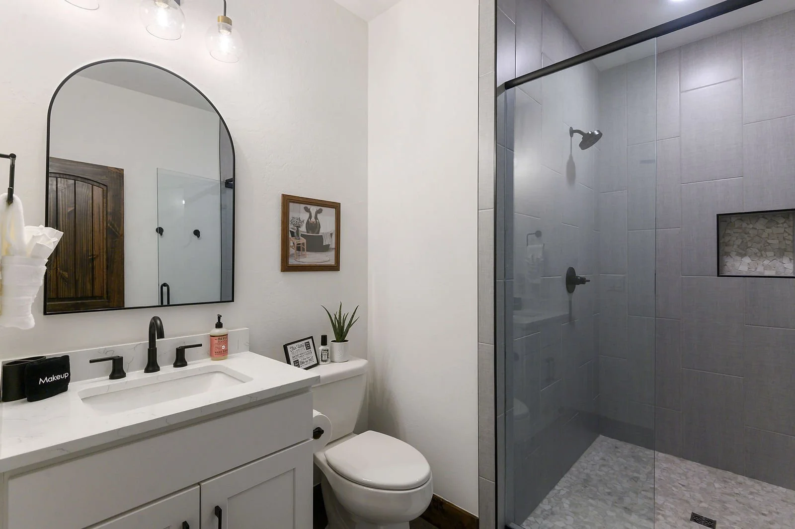 Modern bathroom with a white vanity, black fixtures, a large arched mirror, a framed artwork on the wall, a small potted plant, toiletries, and a walk-in shower with gray tiles and a glass door.