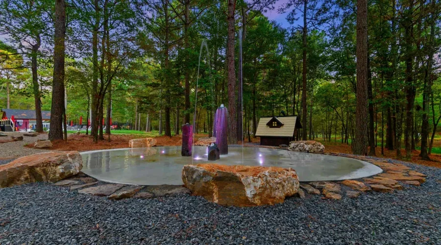 A circular outdoor hot tub surrounded by rocks and trees in a forested area, with two tall water fountains in the center and a small wooden shed nearby.