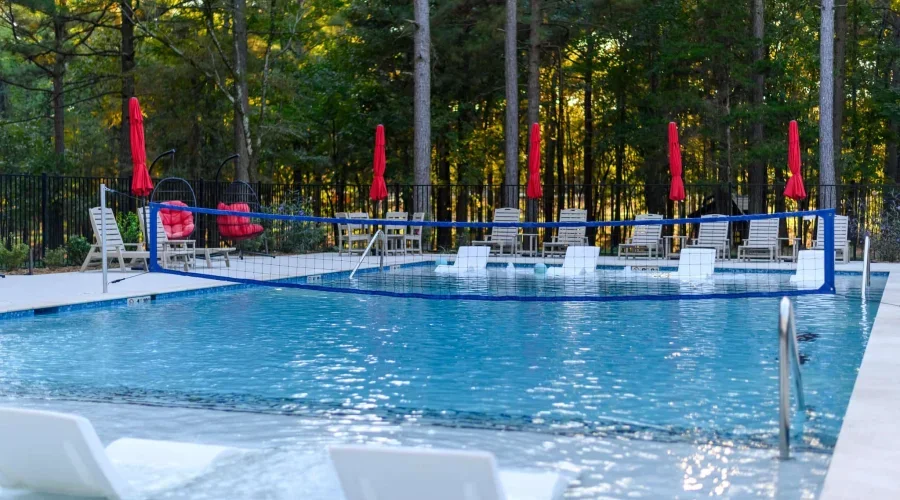 An outdoor swimming pool surrounded by white lounge chairs, red umbrellas, and a black fence, with a wooded area in the background.