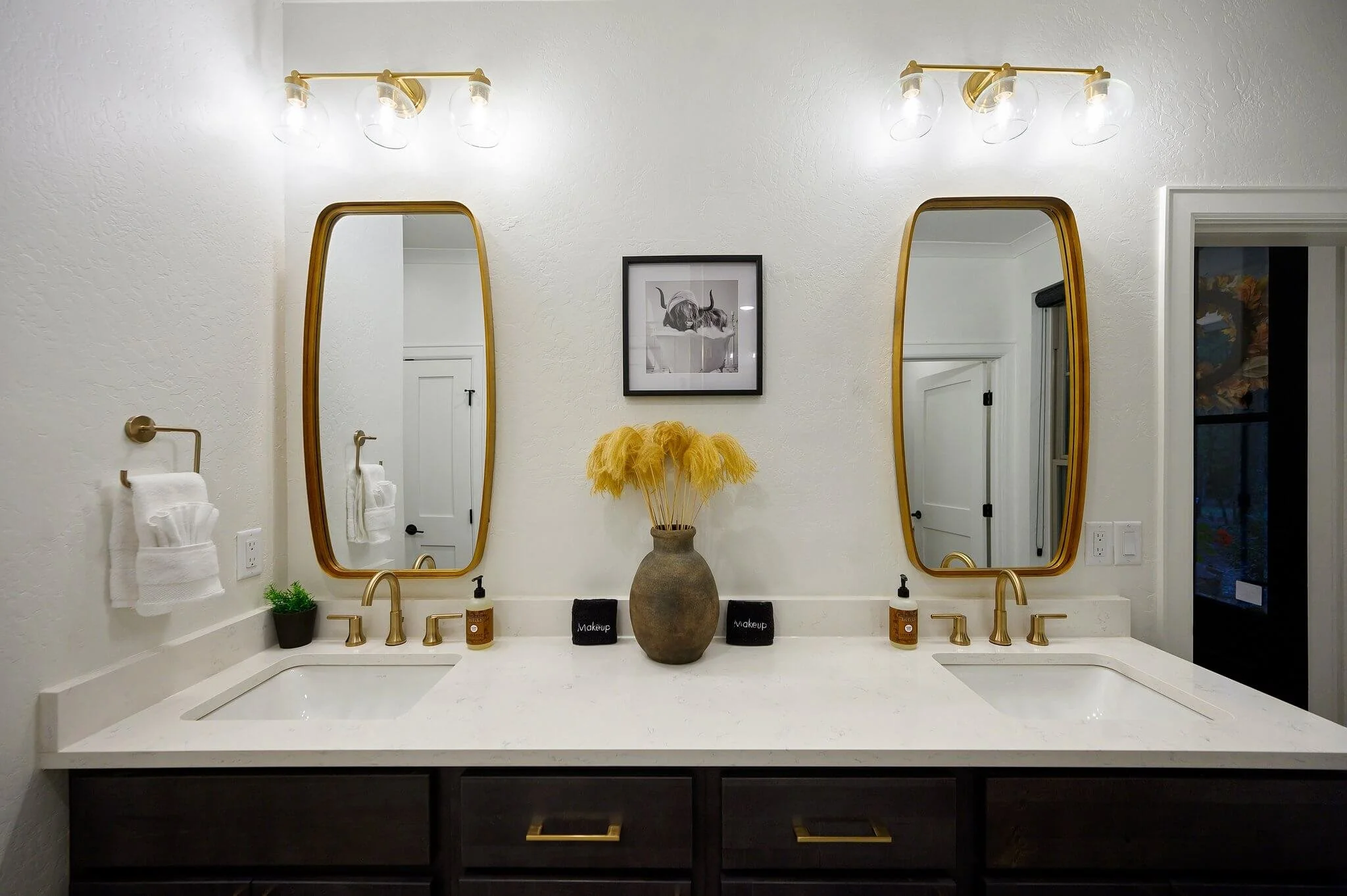 A modern bathroom vanity with a white countertop, two gold sinks, and two gold light fixtures above two large oval mirrors. Decor includes a potted plant, a vase with yellow pampas grass, and small containers labeled 'Makeup' and 'Skincare'.