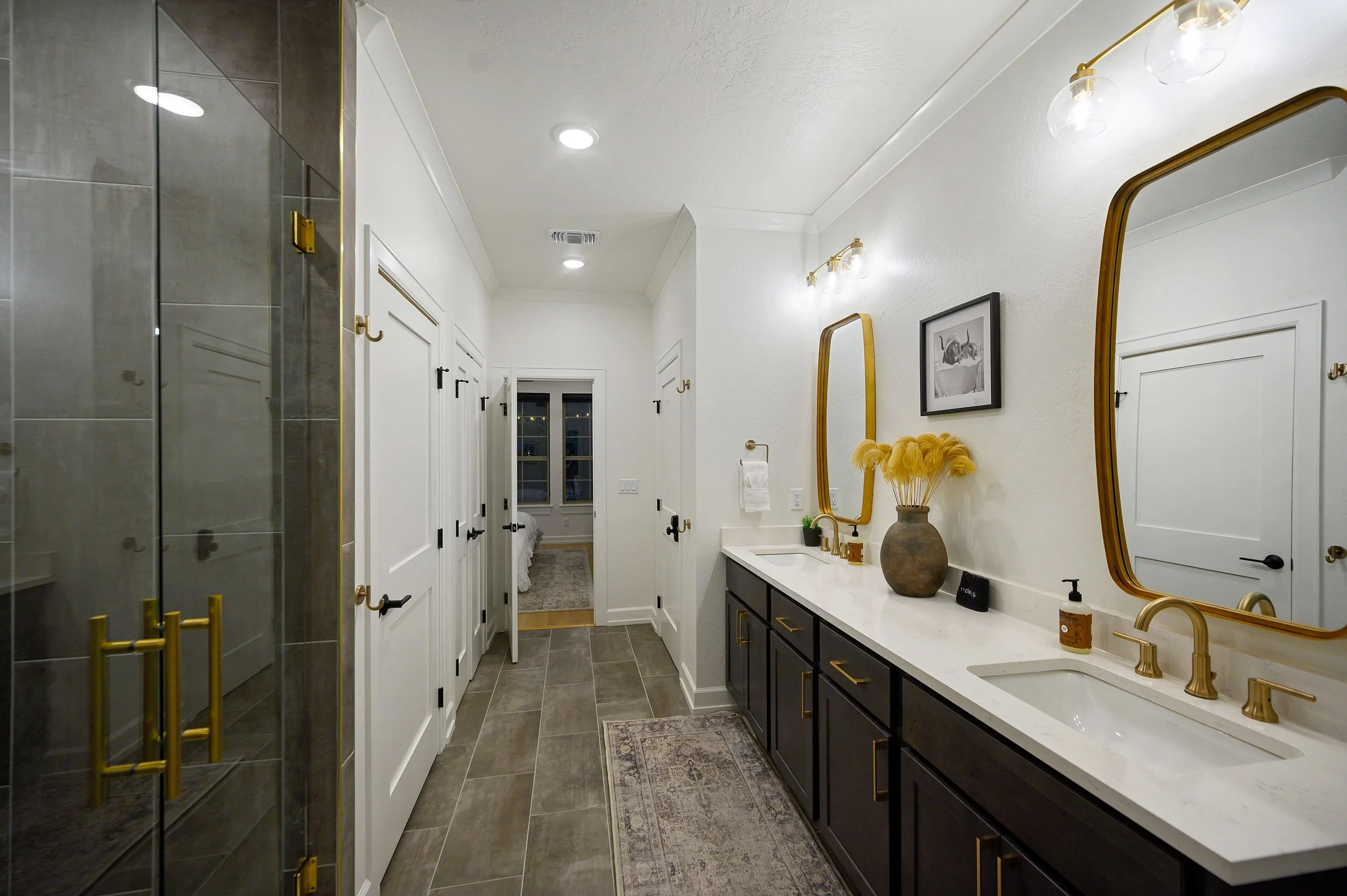 Modern bathroom with a white marble countertop, a gold faucet, a large mirror with a gold frame, black cabinets, decorative items, and a shower with gray tiles and gold fixtures.