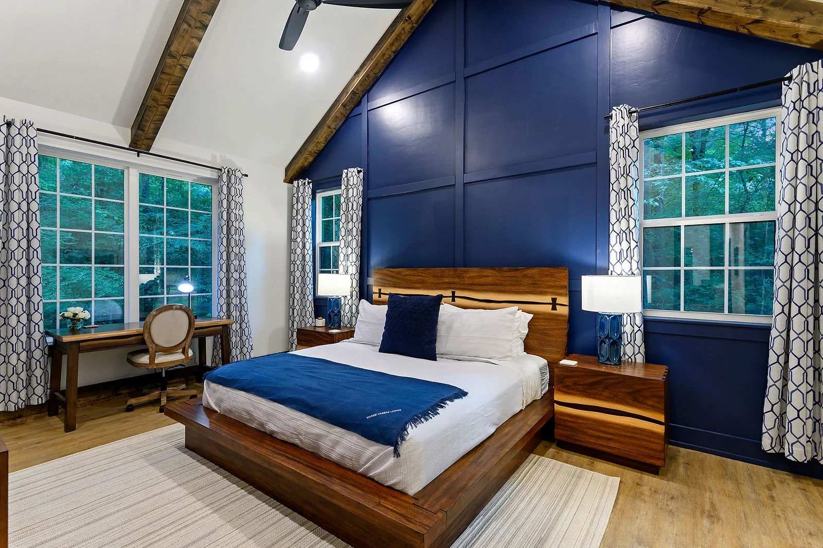 A bedroom with a large wooden bed, blue accent wall, and two windows with patterned curtains. There are two nightstands with lamps, a wooden desk with a chair, and a ceiling fan.