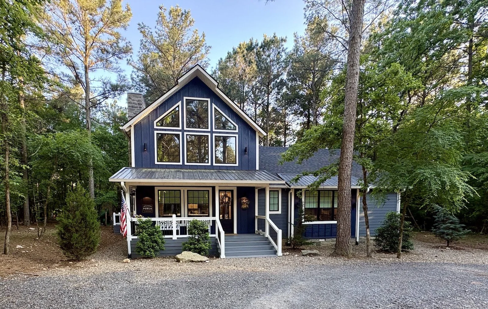A blue two-story house with large windows, a small front porch with white railings, surrounded by trees and bushes, under a clear blue sky.