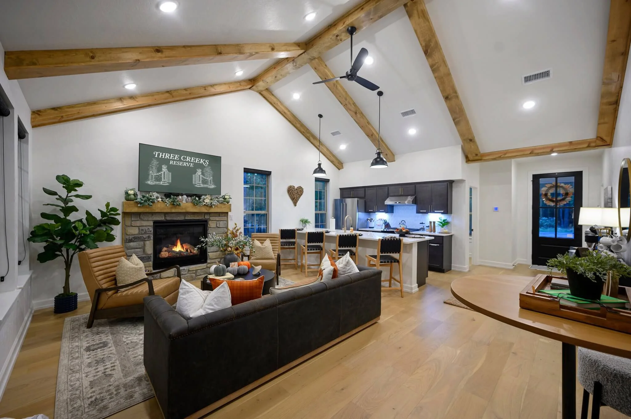 Inside a modern living room with a vaulted ceiling featuring wooden beams, a fireplace with decorations, a ceiling fan, a sofa, armchairs, and a kitchen area in the background.