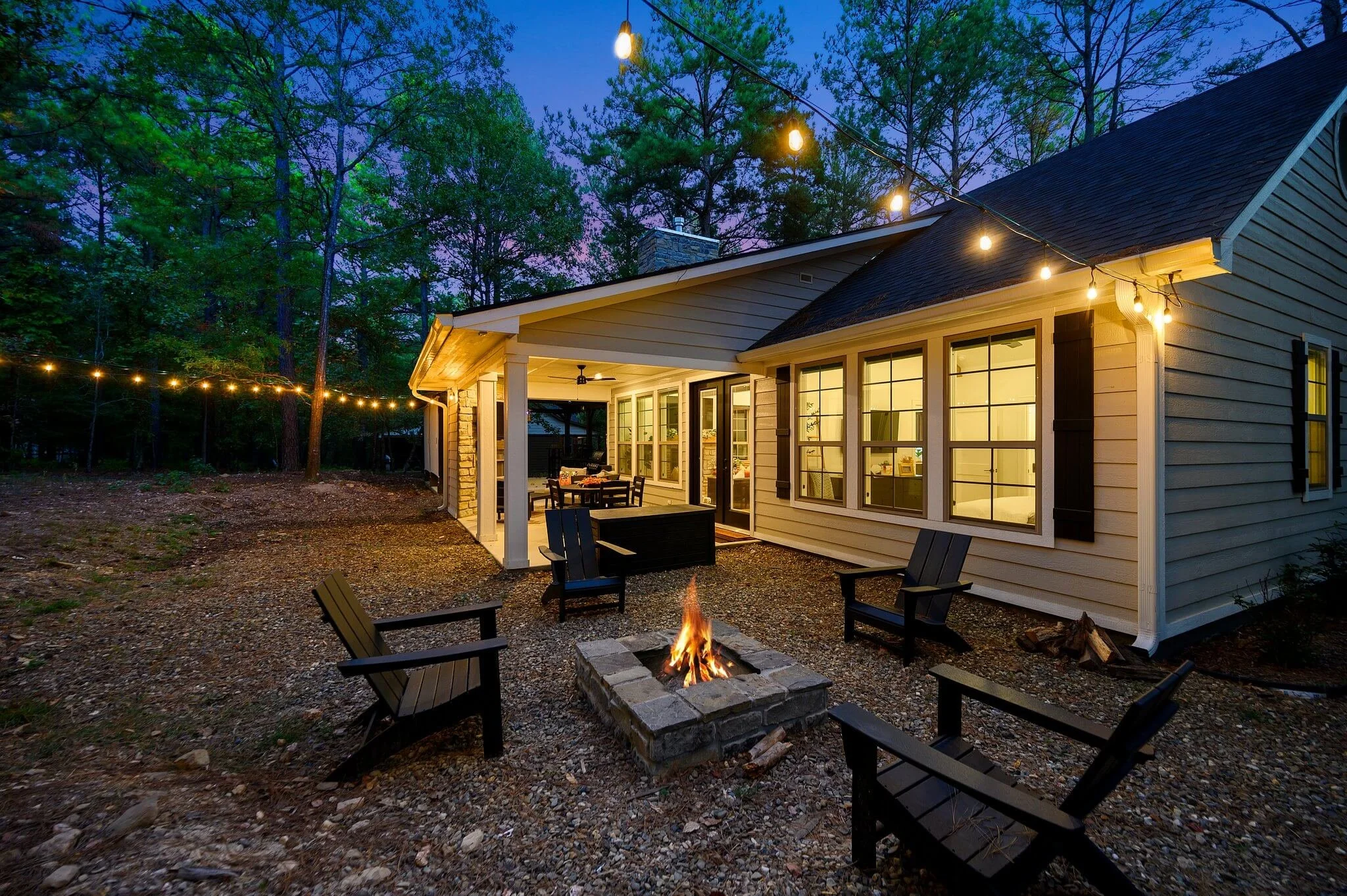 A backyard patio at dusk with string lights, a fire pit, outdoor furniture, and a house with large windows and a covered porch, surrounded by trees.