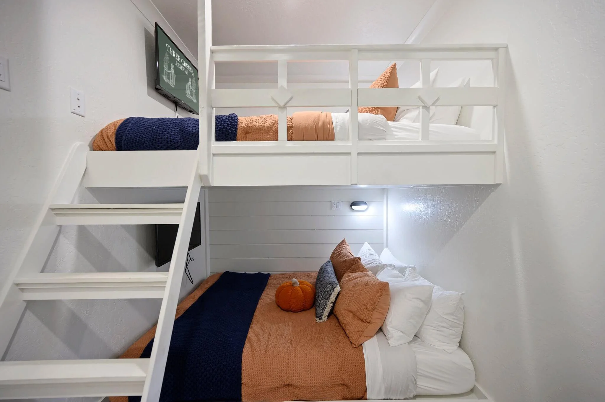 A white bunk bed with a lower bed dressed in orange and white bedding, decorated with pillows and a plush pumpkin, and an upper bed with white bedding and orange pillows. A small flat-screen TV is mounted on the wall near the bed, and the room has wh