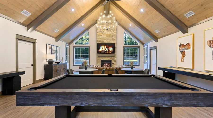 A spacious living room with a vaulted wooden ceiling, a central stone fireplace with a mounted TV above, a game table in the foreground, and seating arranged around the fireplace, with large windows on both sides.