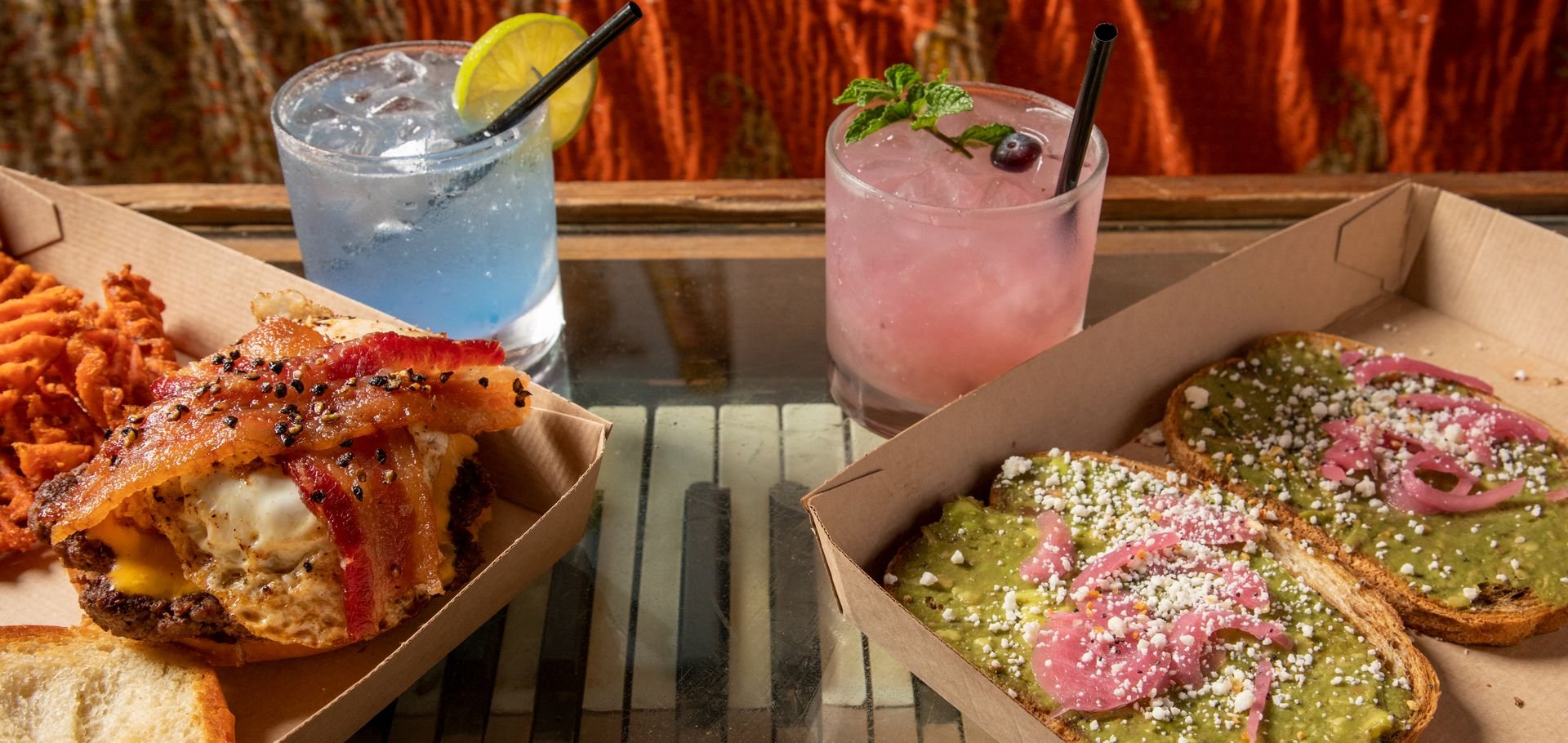 Food and drinks on a table, including a cheeseburger with bacon and black pepper, a box of avocado toast topped with radish and cheese, and two colorful cocktails with lemon and mint garnishes.
