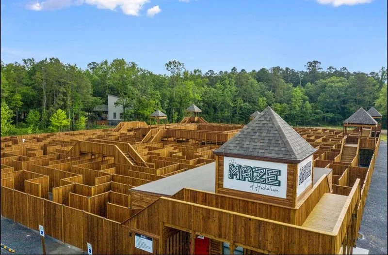 A large wooden maze with pathways and dead ends, surrounded by trees and blue sky, with signs at the entrance.