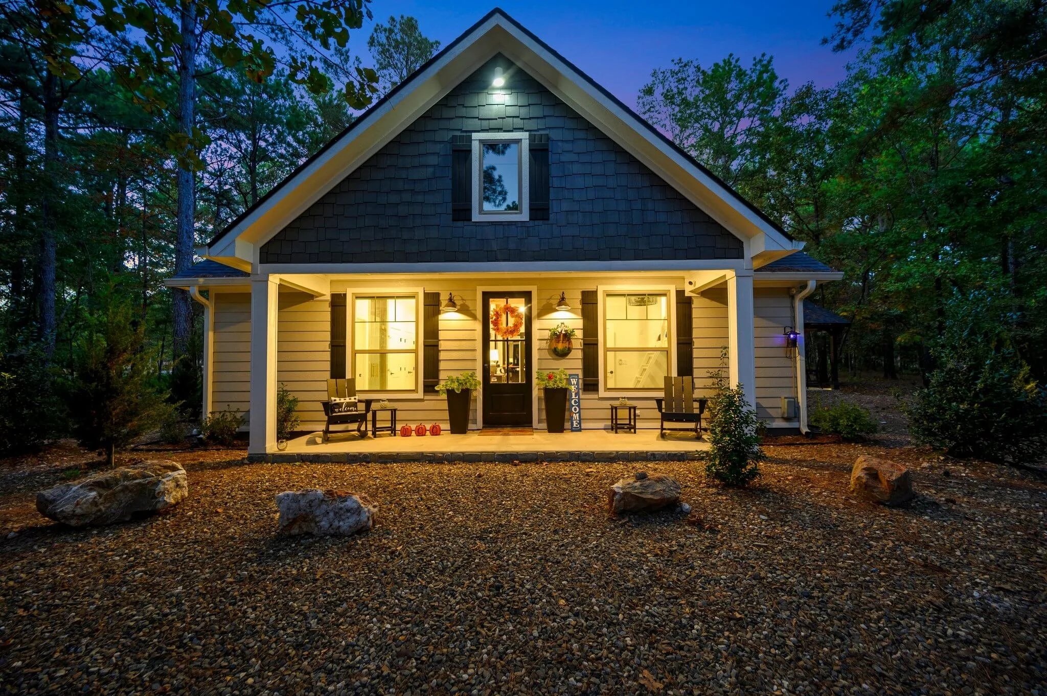 A cozy house with warm lighting on the porch, decorated with fall-themed items, surrounded by trees in a wooded area at dusk.